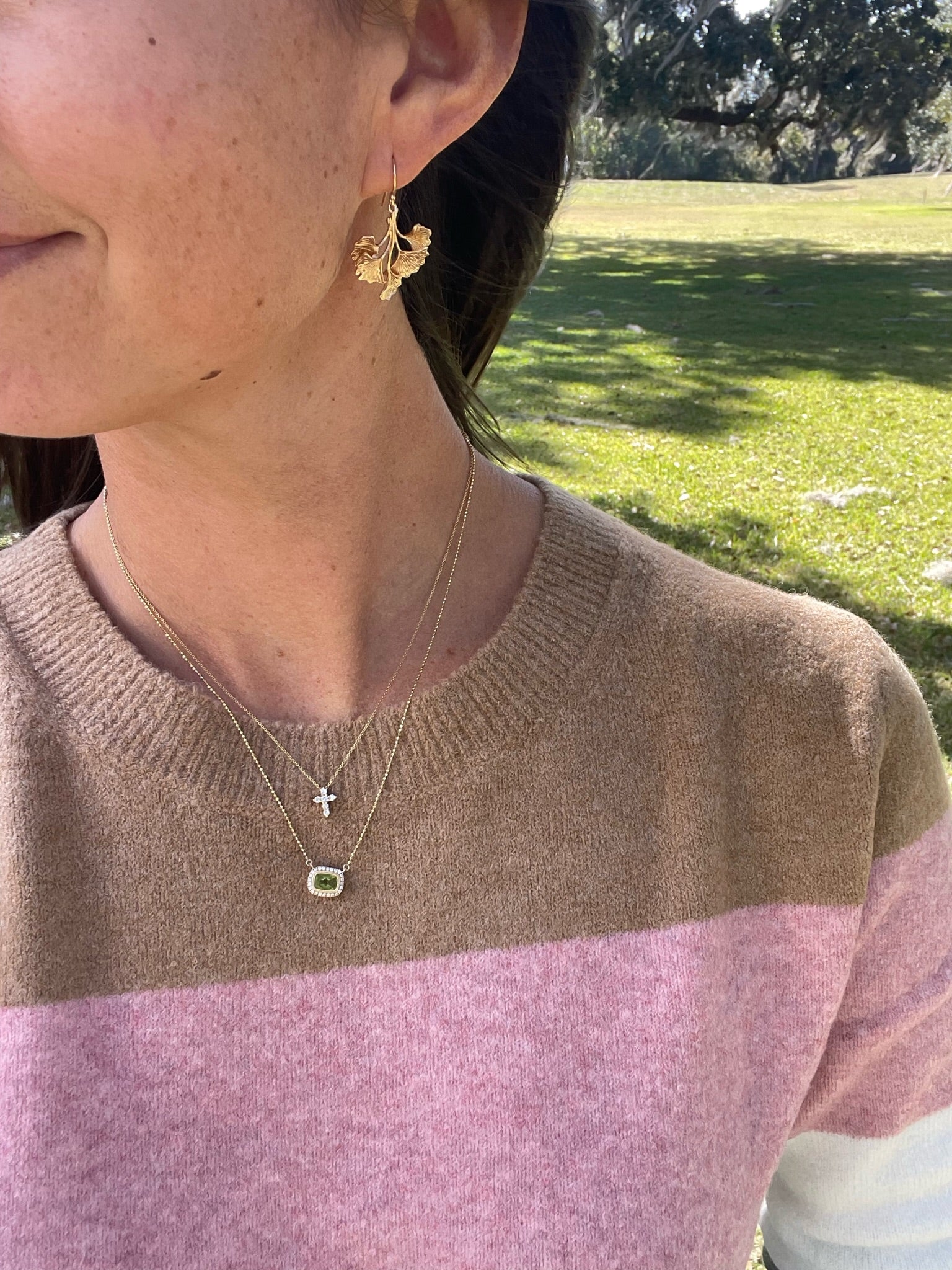 Person wearing earrings, necklace with a cross pendant and green stone, standing outdoors.