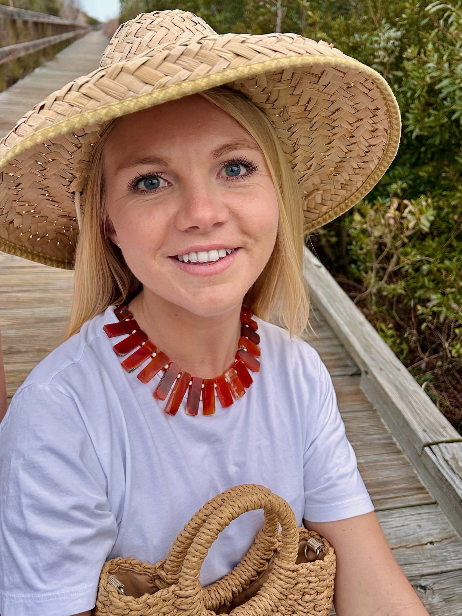 Woman wearing a straw hat and white shirt with a red necklace, standing on a beach boardwalk